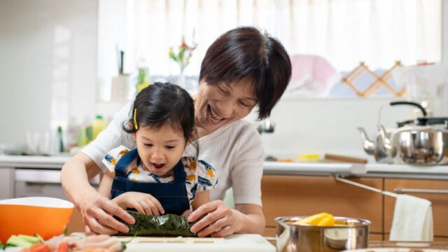 An Asian grandmother holds her young grandchild in her lap as they cook together in the kitchen.
