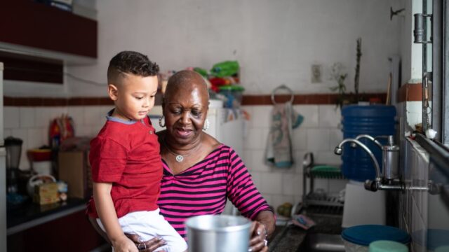 A Black grandmother holds her grandson as she cooks over a large pot in the kitchen.