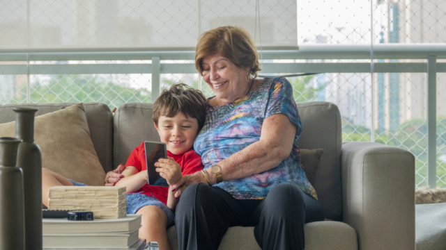 A white woman and her grandson smile as they sit on a couch and look at a smartphone together
