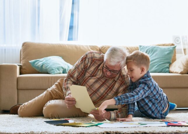 A white grandfather and young grandson sit on the floor and look at papers together.