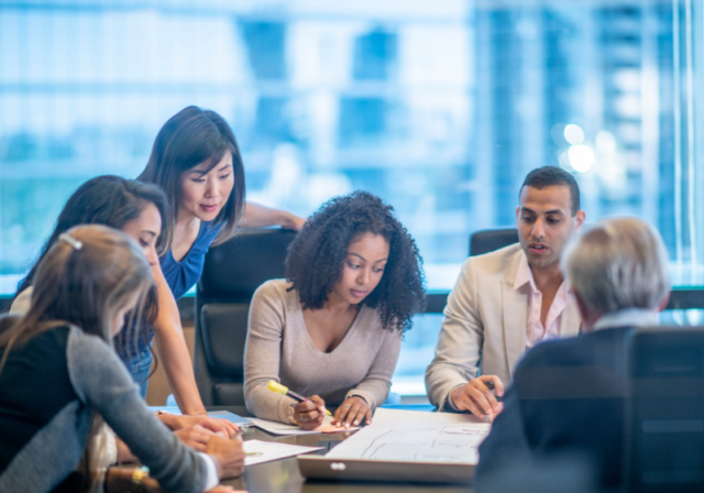 A diverse group of professionals sit around a table and work on papers together.