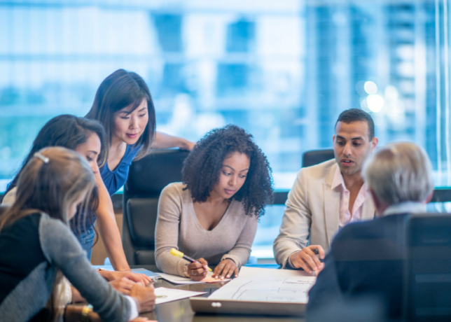 A diverse group of professionals sit around a table and work on papers together.