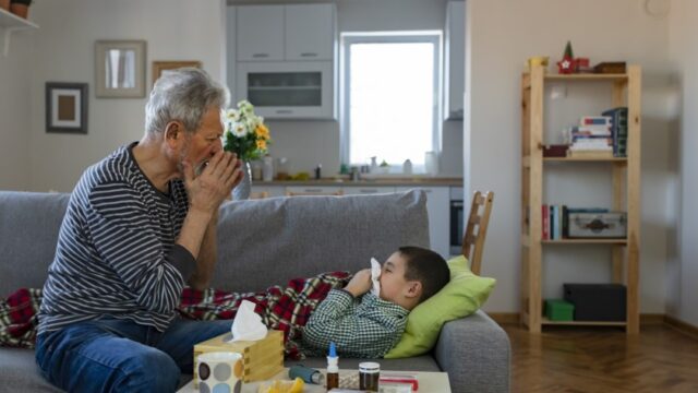 A white grandfather models blowing his nose for his grandson, who is sick and lying on the couch