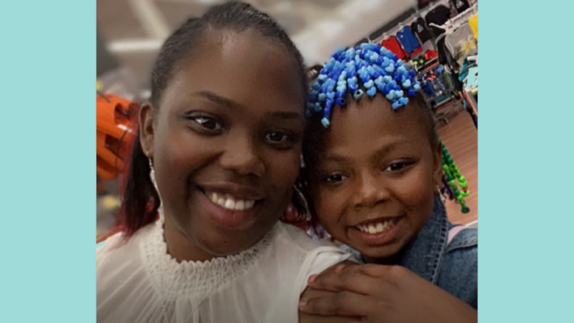 Santana Lee smiles at the camera and her daughter holds on to her shoulder and smiles at the camera alongside her. Her daughter has blue and green beads in her hair.