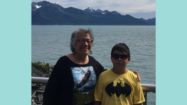 Rosalie Tallbull has her arm around her grandson as both smile for the camera in front of a water and mountain vista