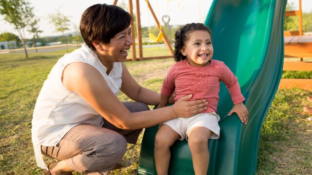 A Latina grandmother plays with her young granddaughter, who is smiling at the bottom of a slide