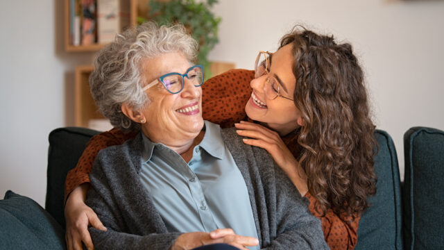 A white grandmother sits on a couch and smiles at her teenage or young adult granddaughter, who is leaning over the back of the couch, embracing her grandmother, and smiling at her.
