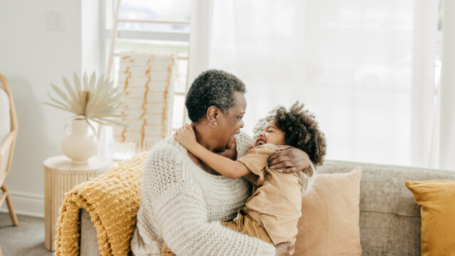 A Black grandmother sits and holds her young grandchild on the couch.