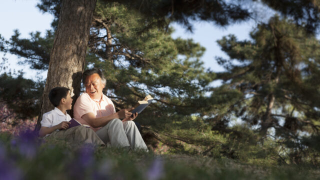 A grandfather and grandson of Asian descent sit under a large evergreen tree and smile at each other. Each has a book in his hand.