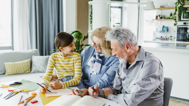 A white granddaughter, grandmother, and grandfather sit at a table together and smile as they color. The child is in the grandmother's lap and they are smiling at each other