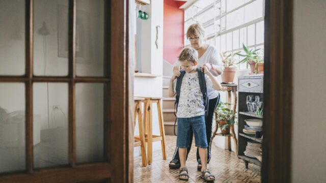 A white grandmother helps her grandson put his school backpack on