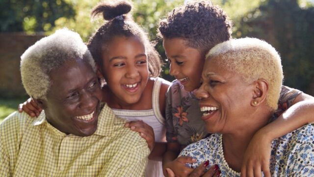 A Black grandmother and grandfather smile at each other and their smiling granddaughter and grandson, who have their arms around their grandparents