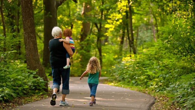 A white grandparent walks with two young grandchildren, holding one in her arms. Their backs are to the camera as they make their way along a paved path in the woods.