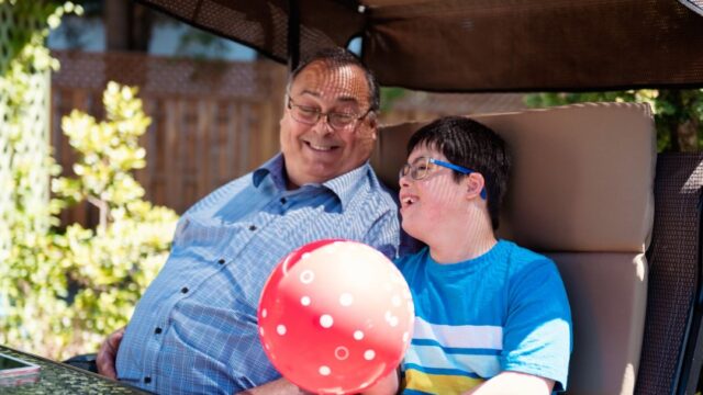 A white grandfather and grandson sit next to each other outside and the grandfather smiles at his smiling grandson, who has a disability and is holding a balloon