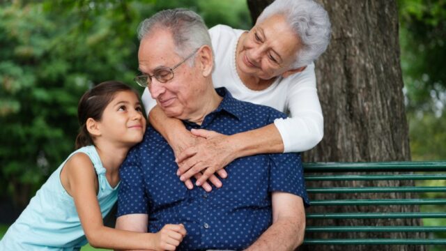 A Latina grandmother leans over behind her husband, who is sitting on a park bench, and clasps her hands on his chest. Both smile at their granddaughter, who is holding her grandfather's arm and smiling at him.