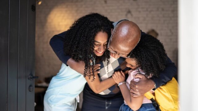 A Black grandfather bends over as he hugs his two granddaughters, one of whom appears to be an adolescent. The older granddaughter's face is partially visible, and she is smiling.