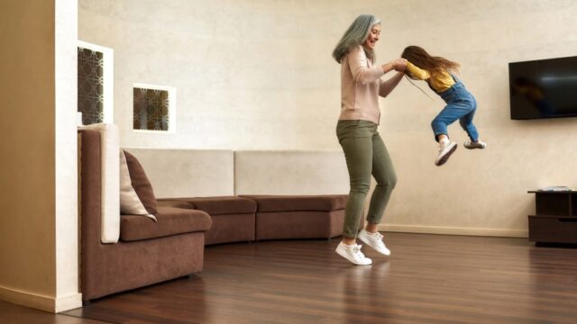 A white grandmother holds her young granddaughter’s hands as the girl jumps in their living room.