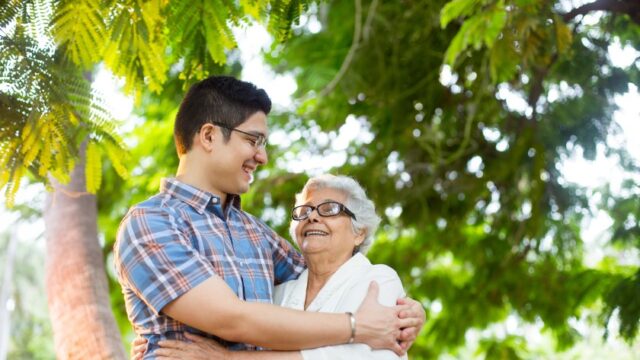 A Latina grandmother and her teenage/young adult grandson smile at each other as they stand outside with their arms around each other.