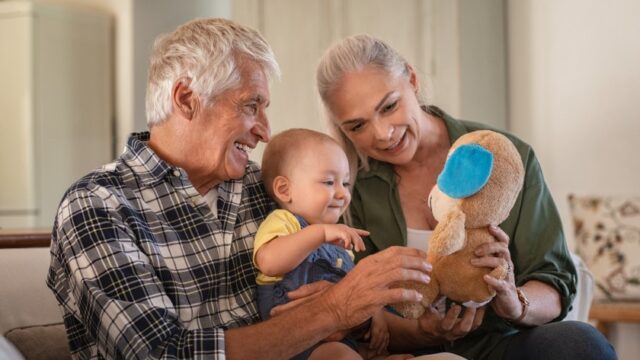 Older adult couple smiling and showing a baby sitting in their lap a stuffed animal.