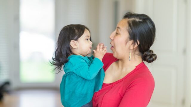 A Native woman holds her young relative in her arms and smiles as the young child claps