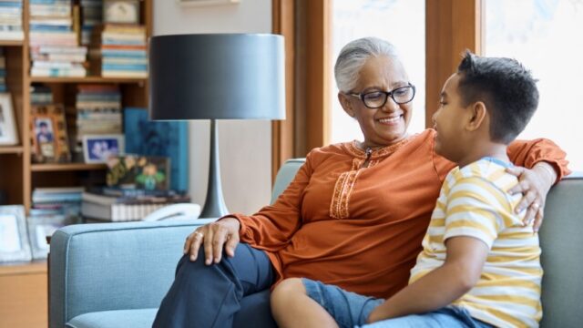 An Indian grandmother and her elementary-aged grandson sit on a couch together and look at each other. The grandmother has her arm around her grandson.