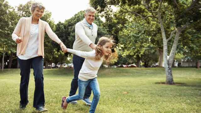 An elementary-aged girl leads her smiling grandparents by the hands through a field. The family is white.