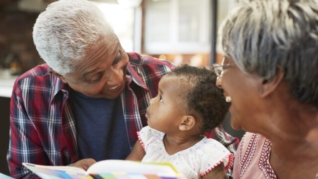 A grandfather smiles at his baby/toddler grandchild, who is looking at him as they read together. The grandmother looks at the two of them with a big smile. The family is Black.