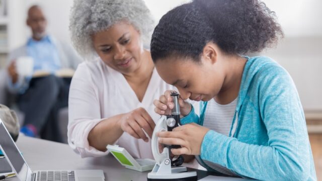 A Black grandmother helps her granddaughter use a microscope. The grandfather is sitting on the couch in the background reading and holding a mug