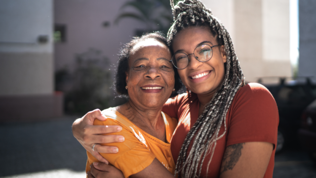 A Black grandmother and her teenage granddaughter smile at the camera with their arms around each other
