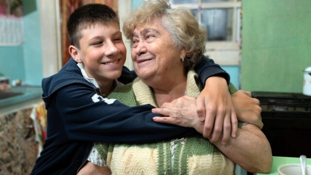 A white grandmother and teenage grandson smile at each other as they sit together. The grandson is hugging his grandmother, and she has her hand on his arm.