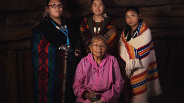A portrait of a grandmother and her three granddaughters inside a Navajo hogan, with the grandmother sitting and the granddaughters standing around her; each of the granddaughters is holding a traditional blanket around herself