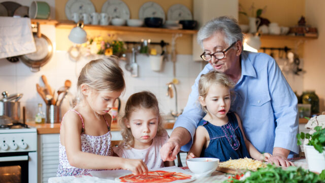 A white grandmother helps her three young granddaughters make homemade pizza the kitchen