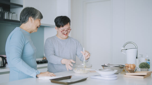 An Asian-American grandmother and her teenage grandson, who appears to have a disability, smile as they cook/bake together