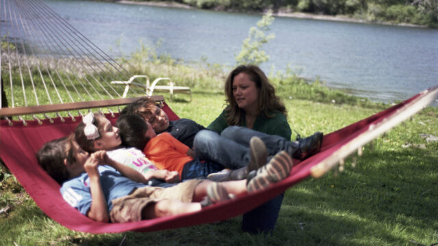 A Native woman kneels by a hammock with four children reclining inside