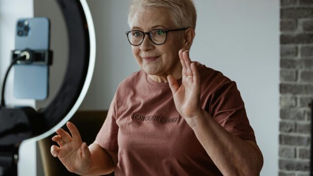 An older woman gestures as she films herself using a smartphone and a ring light