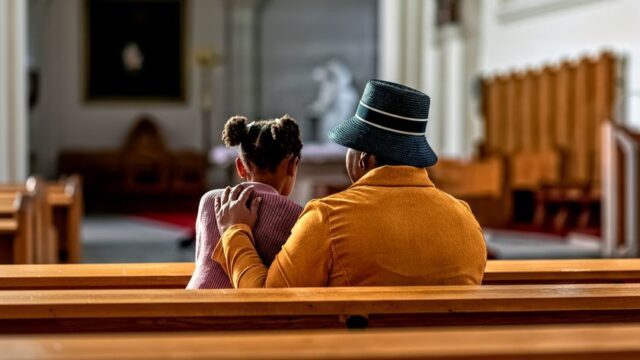 Grandmother and granddaughter sitting inside a church on a wooden bench.